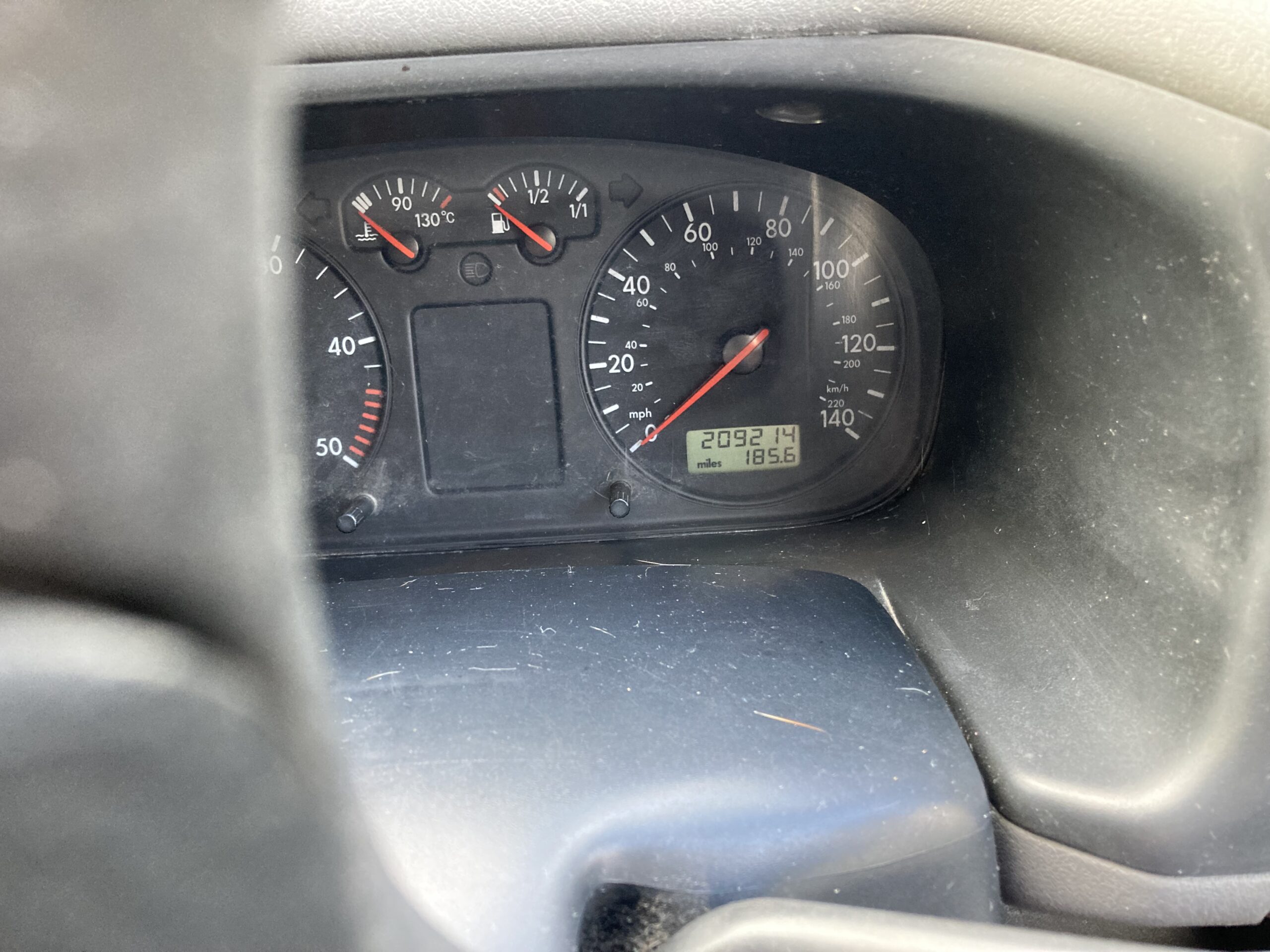 A car dashboard showing the instrument cluster. The speedometer reads approximately 98 km/h, and the odometer reads 209214 km. The fuel gauge is near full, and the temperature gauge is at a safe level. There is some light dust on the dashboard surface.