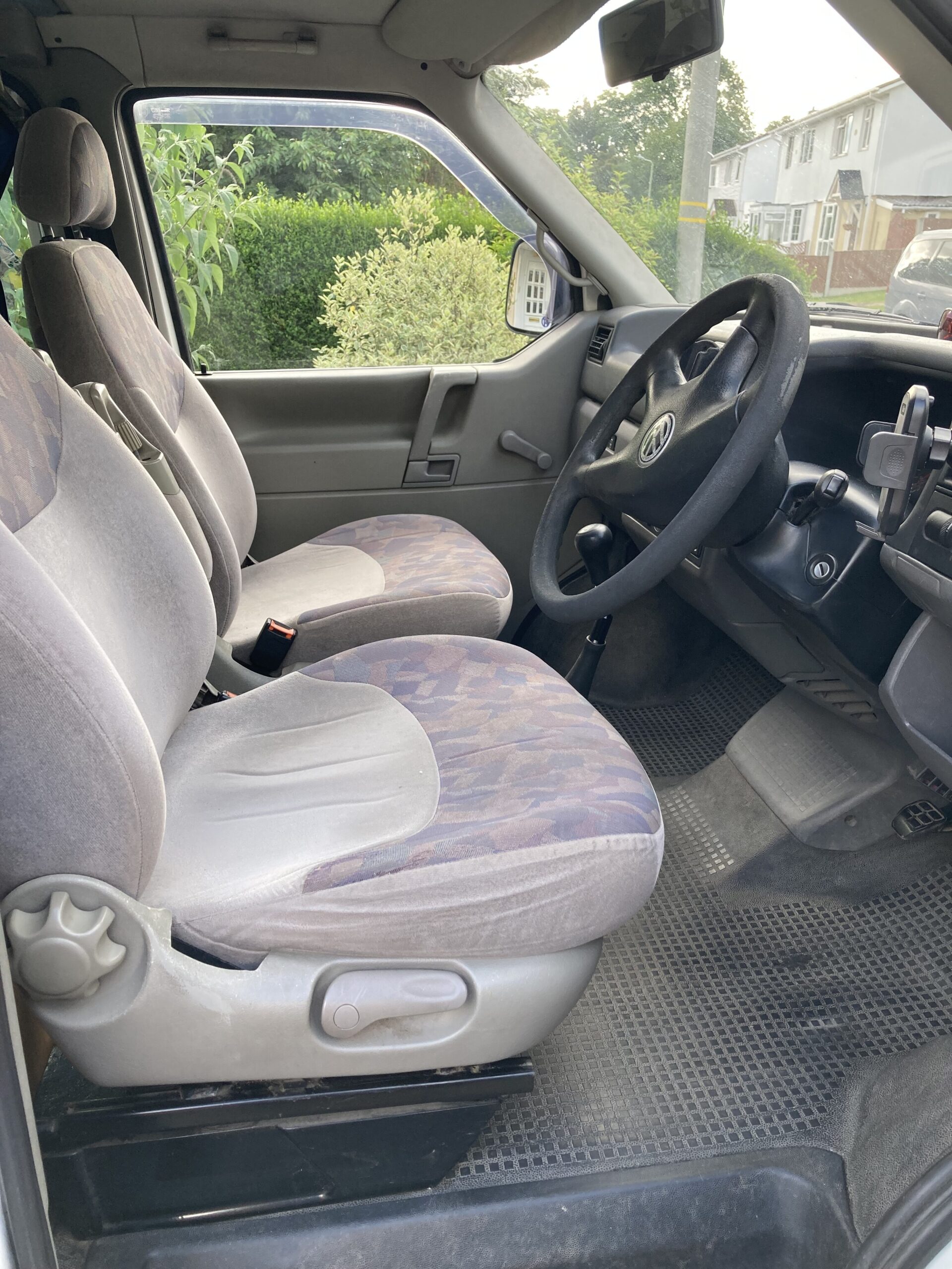 The interior of a car with two front seats upholstered in patterned fabric. The driver's seat has a steering wheel, dashboard, and various controls in view. The passenger seat is beside it. The floor has black rubber mats. Outside through the windows is greenery and part of a residential area.