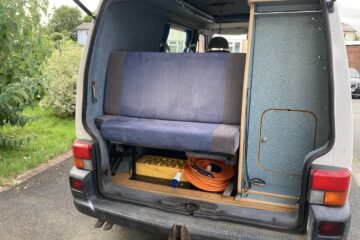 The back of a van is open to reveal a compact living space with a blue upholstered bench, storage compartments, and various items including a yellow crate, orange extension cord, and a pair of shoes. The van is parked on a driveway near greenery and houses.