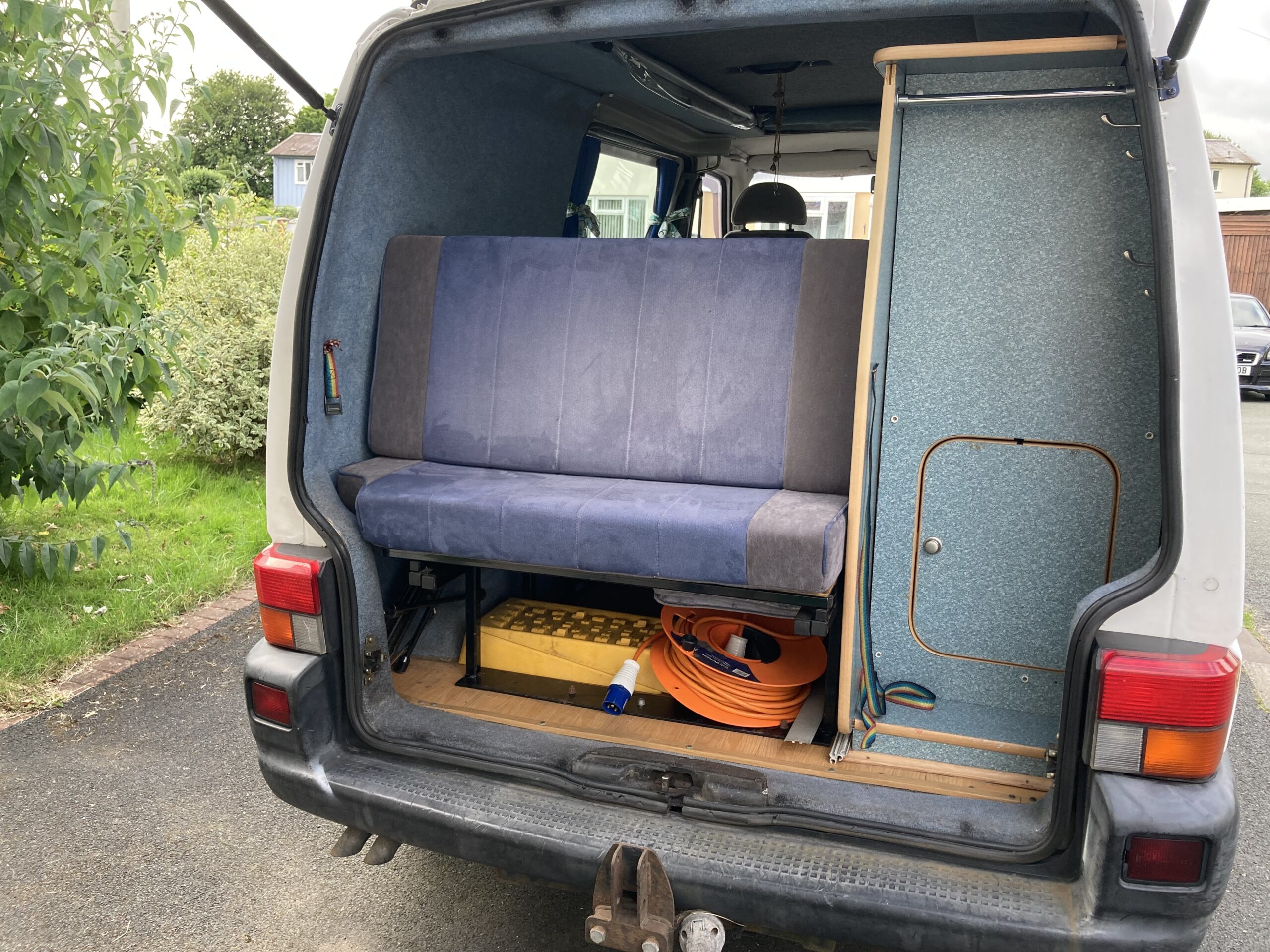 The back of a van is open to reveal a compact living space with a blue upholstered bench, storage compartments, and various items including a yellow crate, orange extension cord, and a pair of shoes. The van is parked on a driveway near greenery and houses.