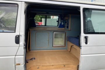 A white van with its side door open reveals a small, neatly organized camper setup. Inside, there are blue cabinets with wooden accents, a small refrigerator, a blue cushioned bench, and blue curtains on the windows. The floor is wood-paneled. A purple leash is on the floor near the open door.