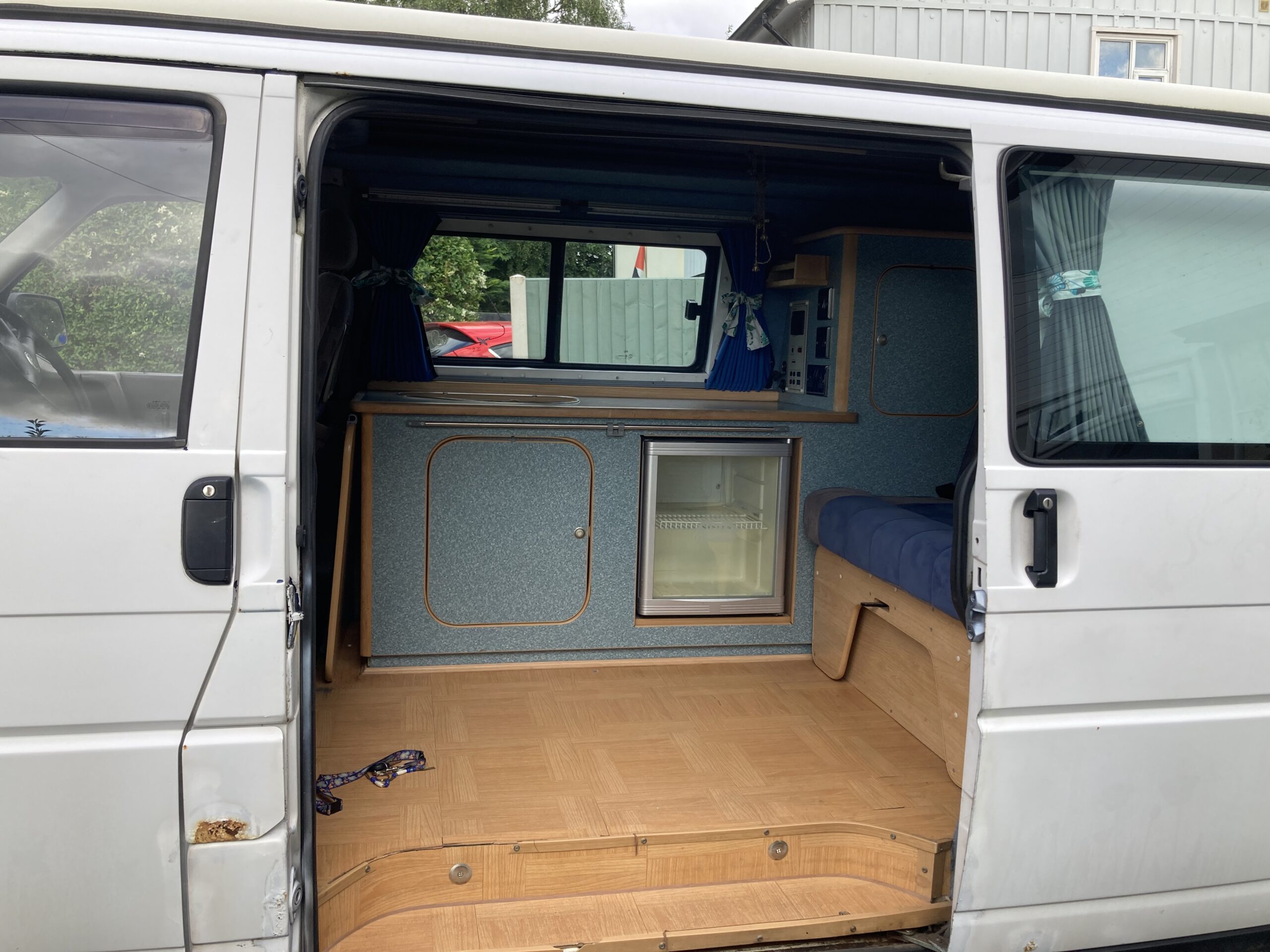 A white van with its side door open reveals a small, neatly organized camper setup. Inside, there are blue cabinets with wooden accents, a small refrigerator, a blue cushioned bench, and blue curtains on the windows. The floor is wood-paneled. A purple leash is on the floor near the open door.
