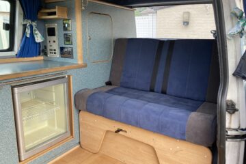 The image shows the interior of a camper van featuring a compact kitchenette with blue and wooden cabinetry. A small refrigerator is built-in below a countertop next to a blue and gray bench seat that doubles as a storage area. Cheerful, patterned curtains hang around the windows.