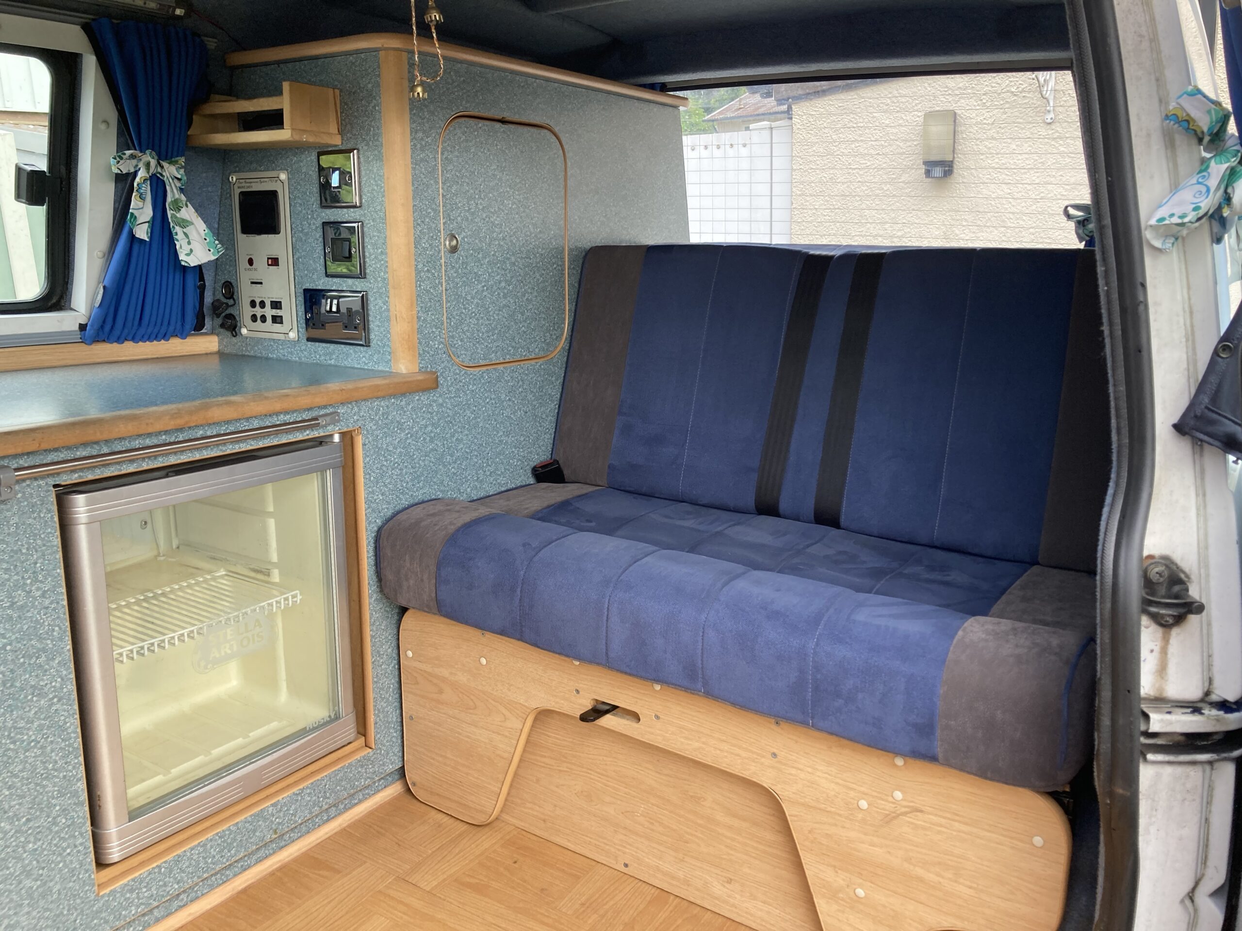 The image shows the interior of a camper van featuring a compact kitchenette with blue and wooden cabinetry. A small refrigerator is built-in below a countertop next to a blue and gray bench seat that doubles as a storage area. Cheerful, patterned curtains hang around the windows.