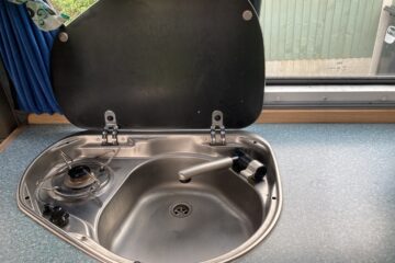 A compact stainless steel sink and single-burner gas stove with a black lid hinged open. The sink has a swivel faucet, and the burner has a control knob. The setup is mounted in a blue-speckled countertop. A window with a blue curtain is above, showing part of a green fence outside.