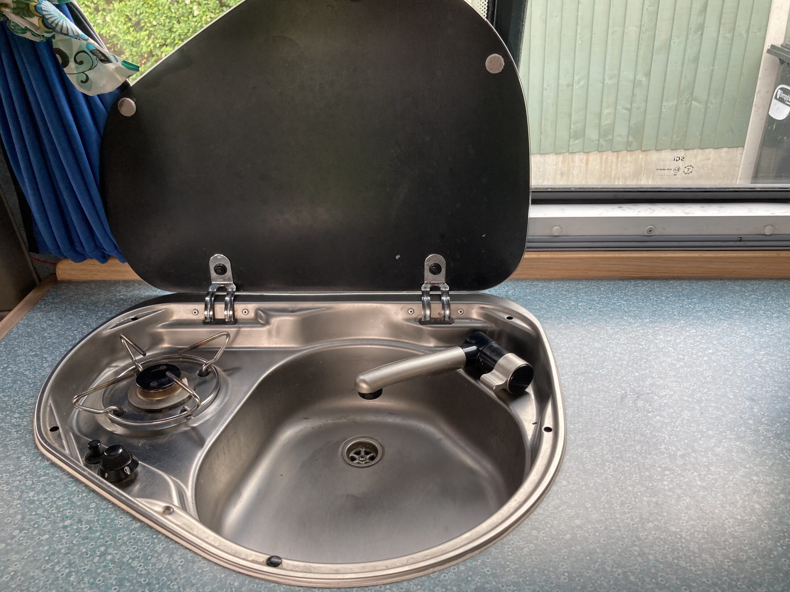 A compact stainless steel sink and single-burner gas stove with a black lid hinged open. The sink has a swivel faucet, and the burner has a control knob. The setup is mounted in a blue-speckled countertop. A window with a blue curtain is above, showing part of a green fence outside.