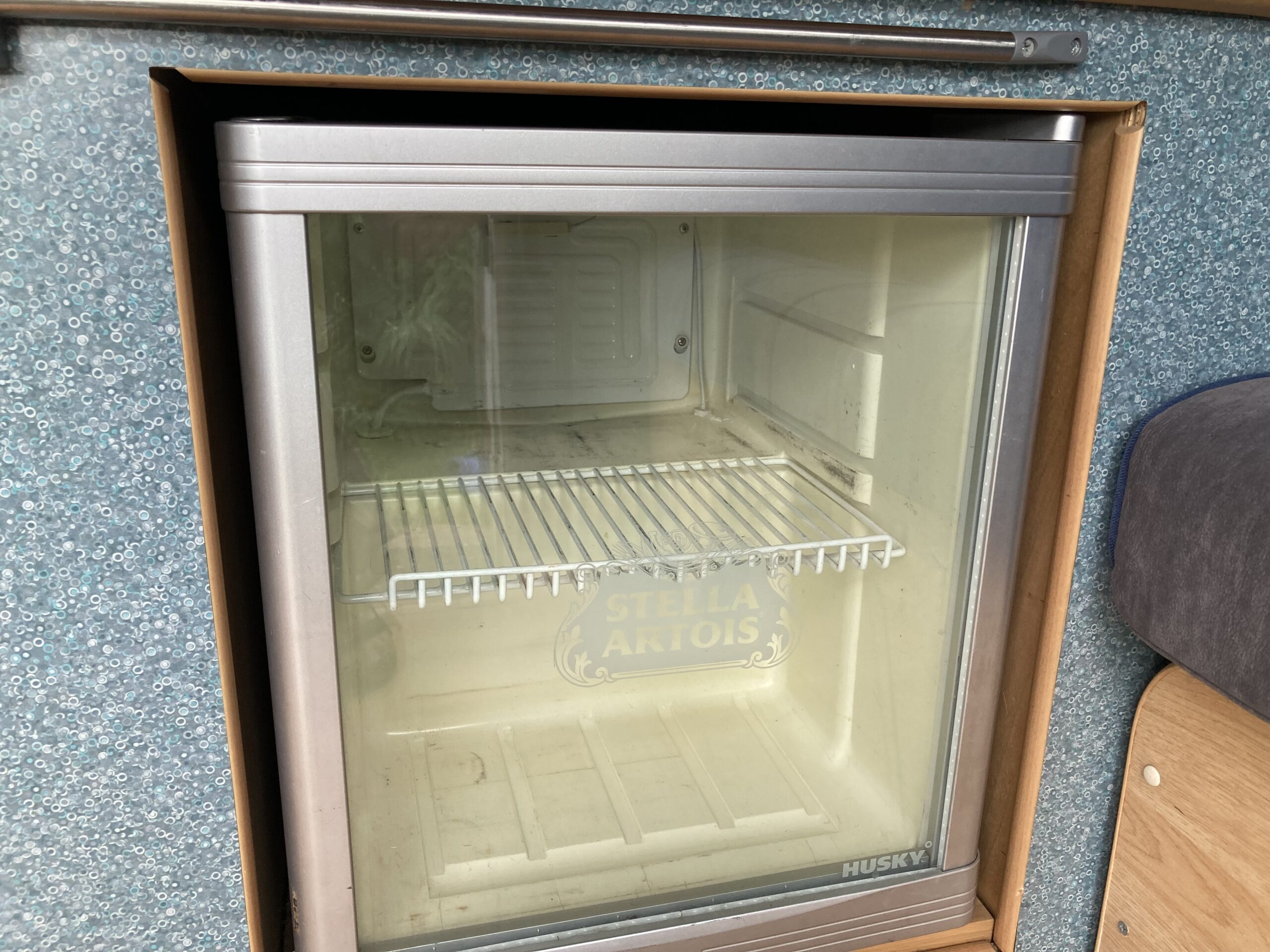 A small, stainless steel Husky mini-fridge with a transparent front door and a wire shelf. The fridge is empty, and the back wall features a faint Stella Artois logo. The exterior is set against a blue, textured surface.