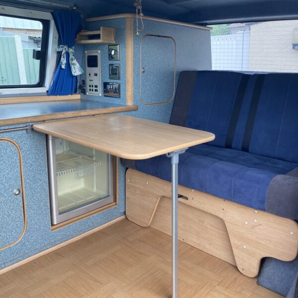 Interior of a camper van featuring a small wooden table attached to the wall, a blue cushioned bench seat, a mini-fridge beneath the table, control panels on the wall, and blue curtains over the window. The floor has a wood grain pattern, and the overall color scheme is blue and wood tones.