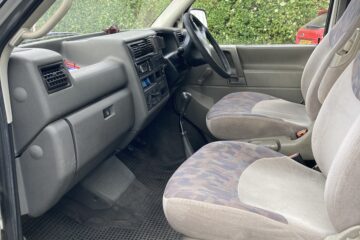 Interior view of a vehicle from the passenger side, showing the two front seats, dashboard, steering wheel, and center console. The seats are upholstered in fabric with a patterned design. The floor is covered with a rubber mat. The windows reveal a green hedge and a red car outside.