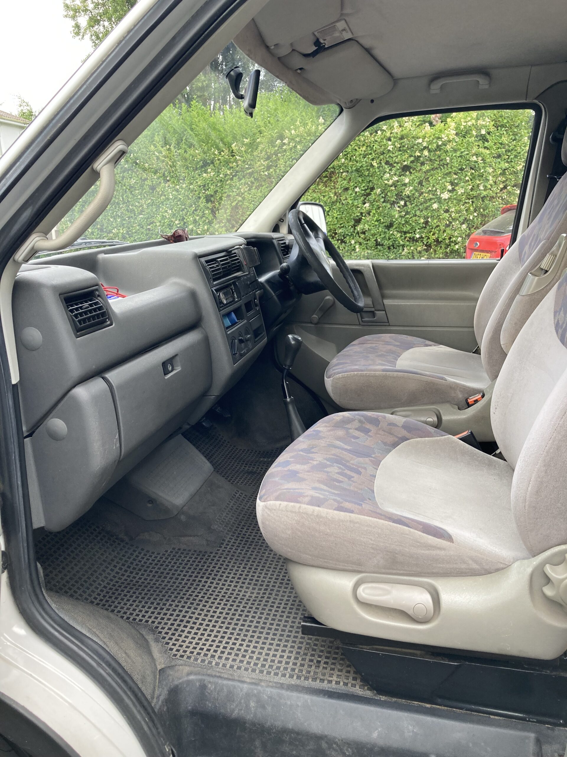 Interior view of a vehicle from the passenger side, showing the two front seats, dashboard, steering wheel, and center console. The seats are upholstered in fabric with a patterned design. The floor is covered with a rubber mat. The windows reveal a green hedge and a red car outside.