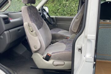 A view of the interior of a vehicle, showing the front passenger and driver seats with a camouflage pattern. The seats are grey with fabric upholstery. The vehicle door is open, revealing a glimpse of the dashboard and steering wheel. Green foliage is visible outside the open door.