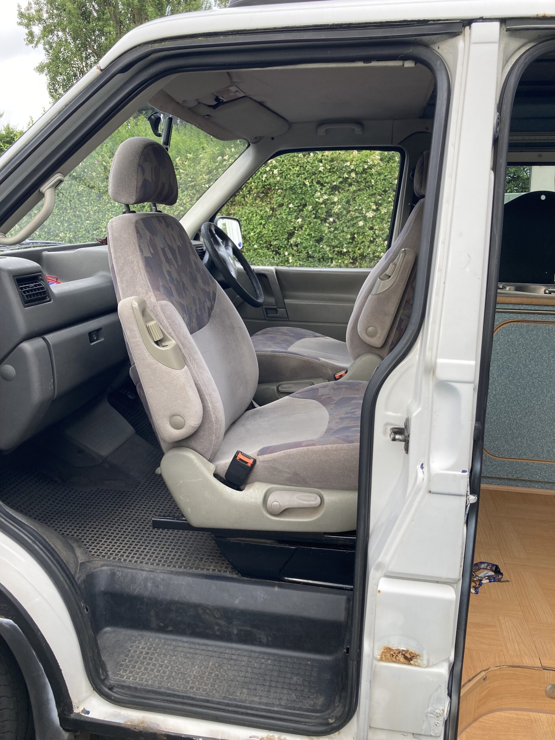 A view of the interior of a vehicle, showing the front passenger and driver seats with a camouflage pattern. The seats are grey with fabric upholstery. The vehicle door is open, revealing a glimpse of the dashboard and steering wheel. Green foliage is visible outside the open door.