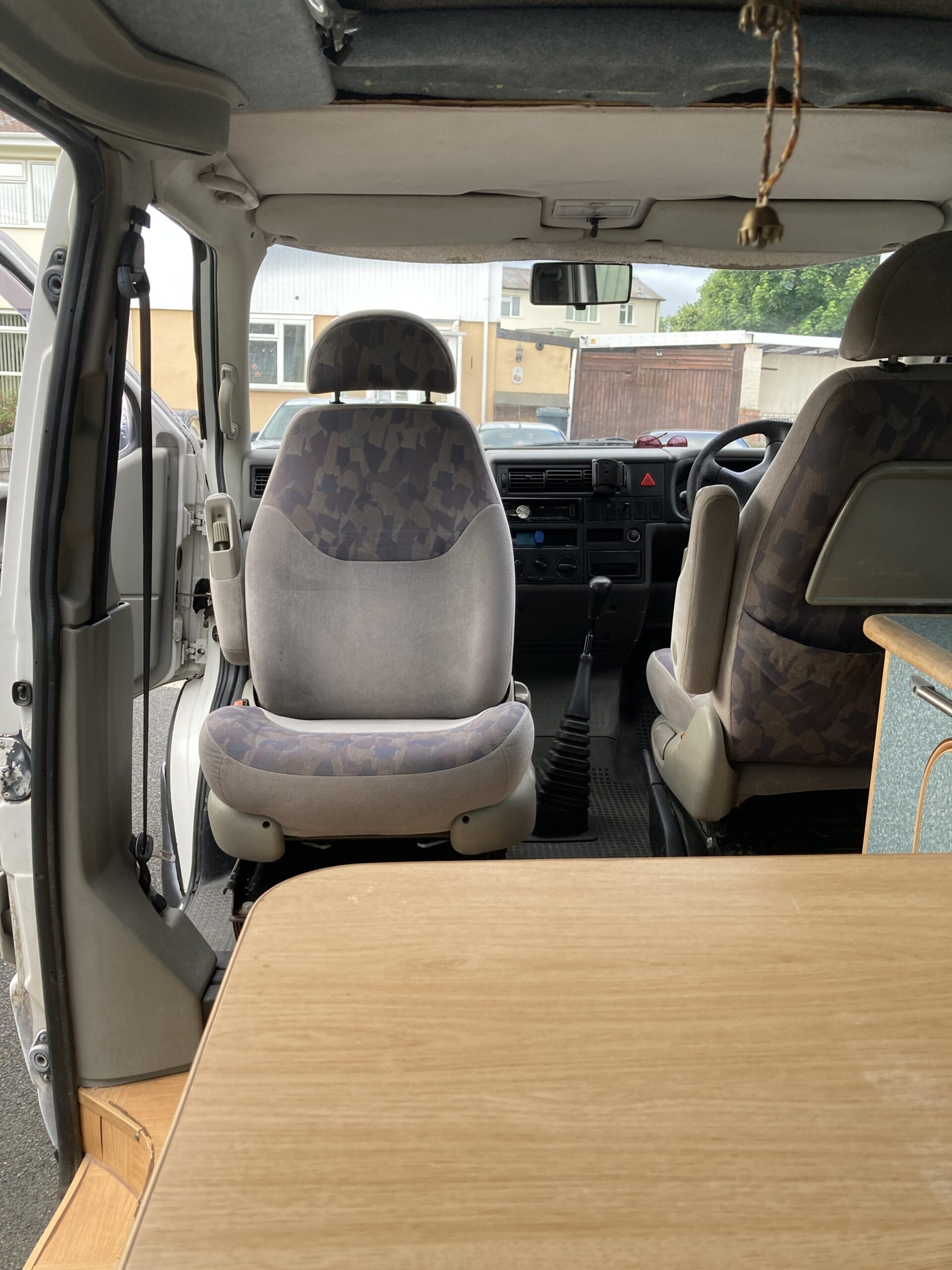 The interior of a camper van with a front passenger seat facing backward. The driverβs seat faces forward, and the dashboard, steering wheel, and gear shift are visible. A wooden table is partially visible in the foreground. The van door is open, showing an outdoor setting.