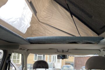 Interior view of a camper van with the pop-top canvas roof extended upward. The front driver and passenger seats are visible, with the back of the seats adorned in a patterned fabric. Outside the vehicle, several cars and a residential building can be seen through the windows.