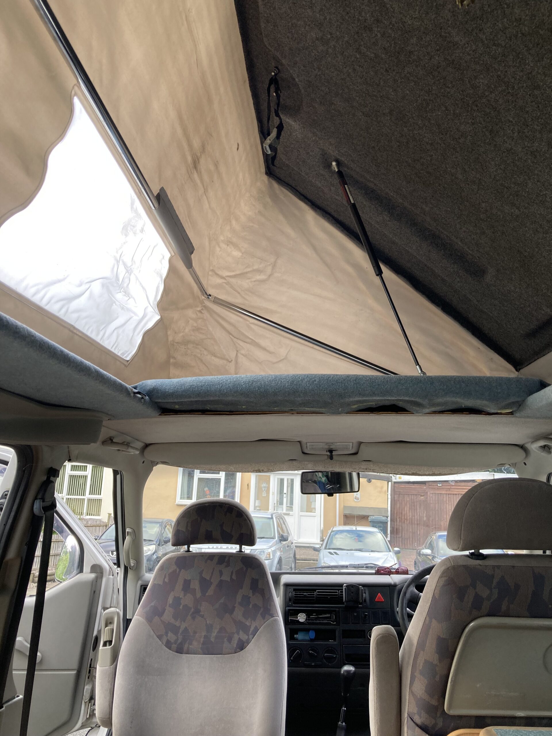 Interior view of a camper van with the pop-top canvas roof extended upward. The front driver and passenger seats are visible, with the back of the seats adorned in a patterned fabric. Outside the vehicle, several cars and a residential building can be seen through the windows.