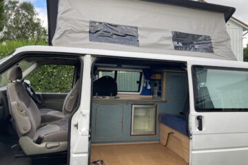 A white campervan with its sliding door open, revealing a compact interior setup. The van's roof is extended upwards, creating more space inside. Inside, there's seating, a small kitchenette with a mini-fridge, and storage cabinets. The van is parked outdoors, near some greenery.