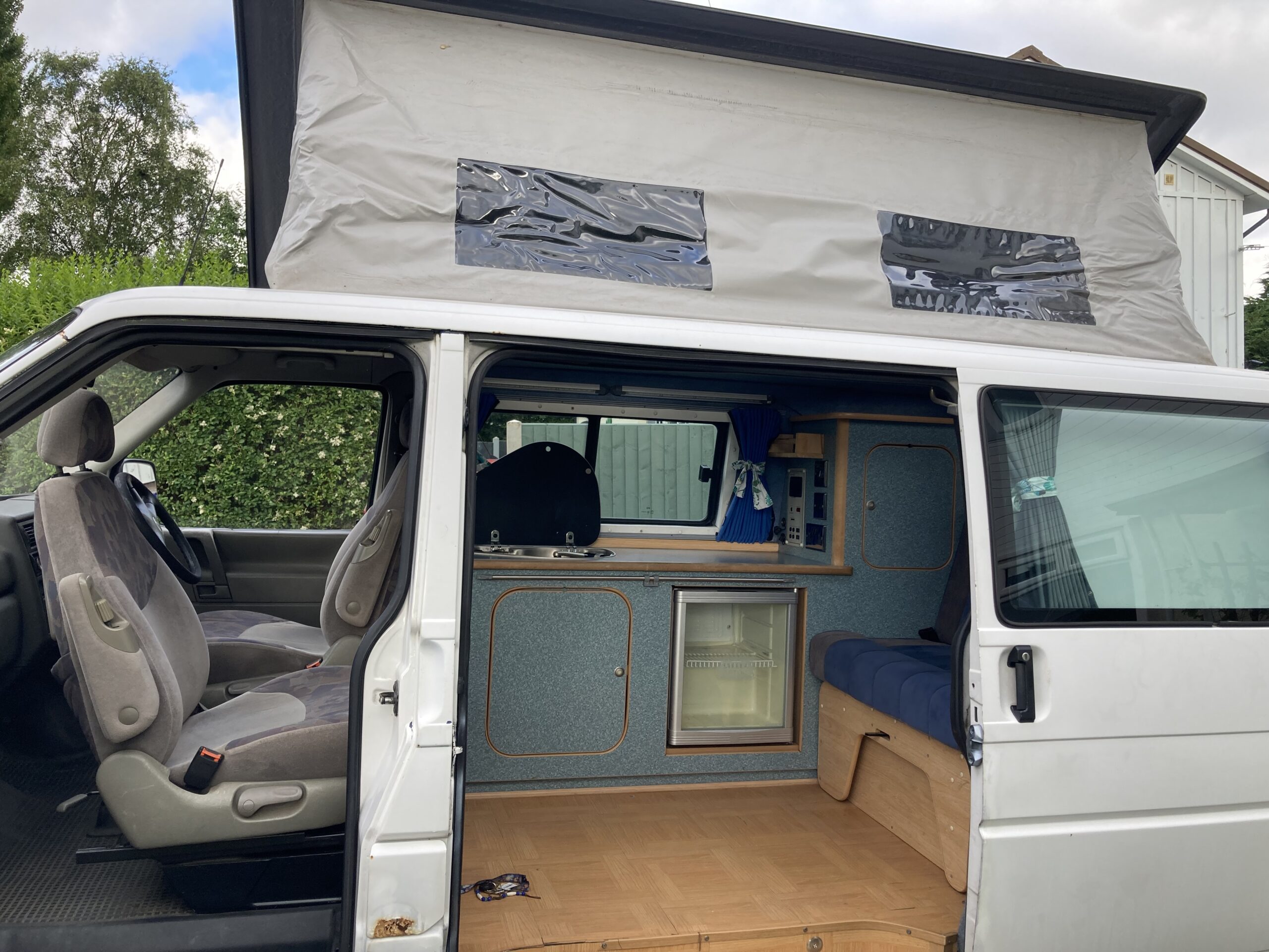 A white campervan with its sliding door open, revealing a compact interior setup. The van's roof is extended upwards, creating more space inside. Inside, there's seating, a small kitchenette with a mini-fridge, and storage cabinets. The van is parked outdoors, near some greenery.