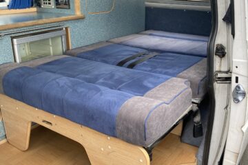 The interior of a camper van is shown with a neatly made bed featuring blue and gray bedding. The wooden frame supports the bed, and there is a small refrigerator and control panel in the background. The van's doors are open, revealing a parquet-style wooden floor and blue curtains on the windows.