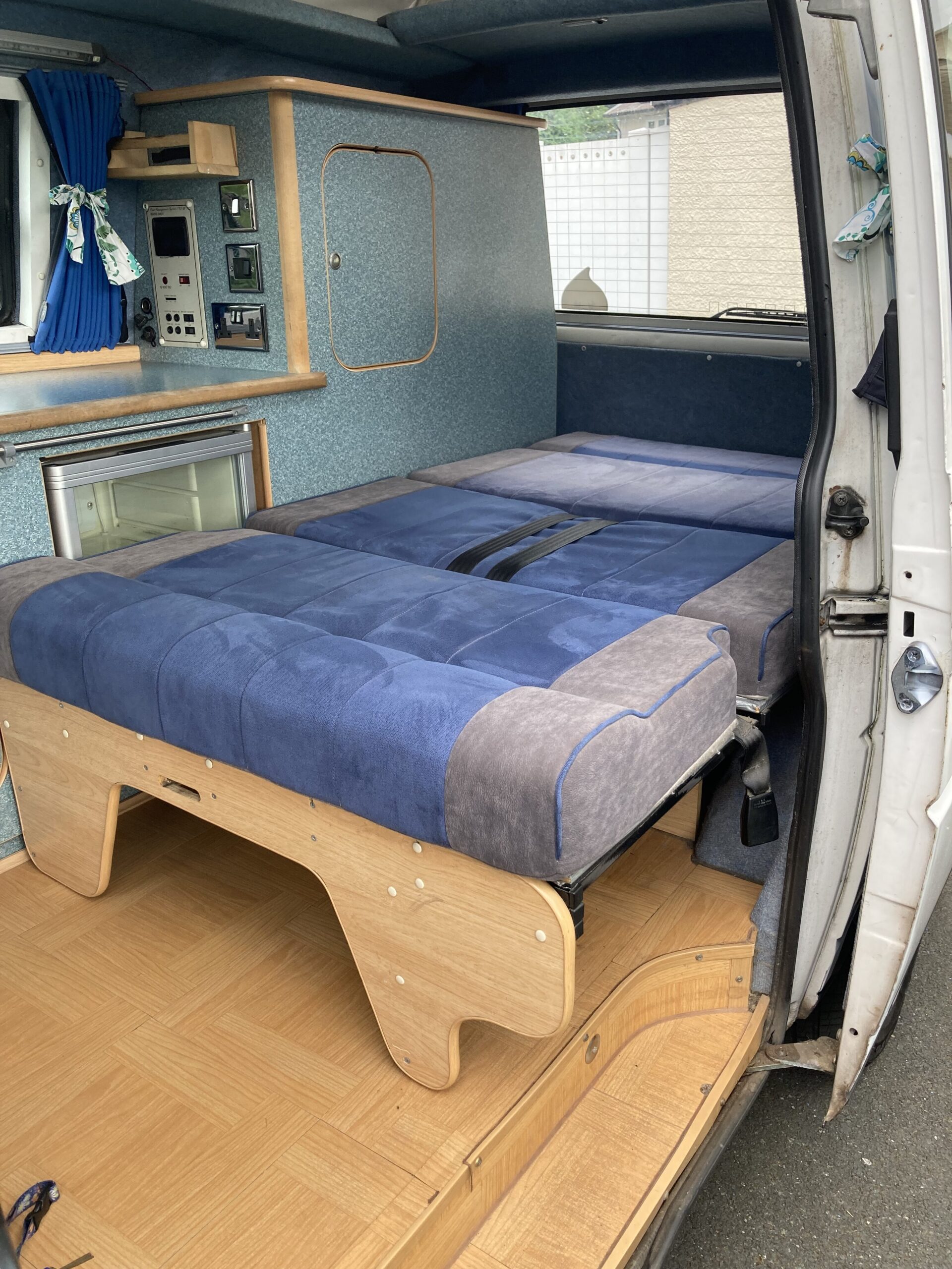 The interior of a camper van is shown with a neatly made bed featuring blue and gray bedding. The wooden frame supports the bed, and there is a small refrigerator and control panel in the background. The van's doors are open, revealing a parquet-style wooden floor and blue curtains on the windows.