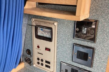 A close-up view of a wall panel in a caravan. The panel features multiple electrical outlets and switches, including UK-style power sockets, a light switch, a 12V DC outlet, and circuit breakers. Above the panel is a wooden rail and a blue curtain. The wall has a textured, light blue finish.