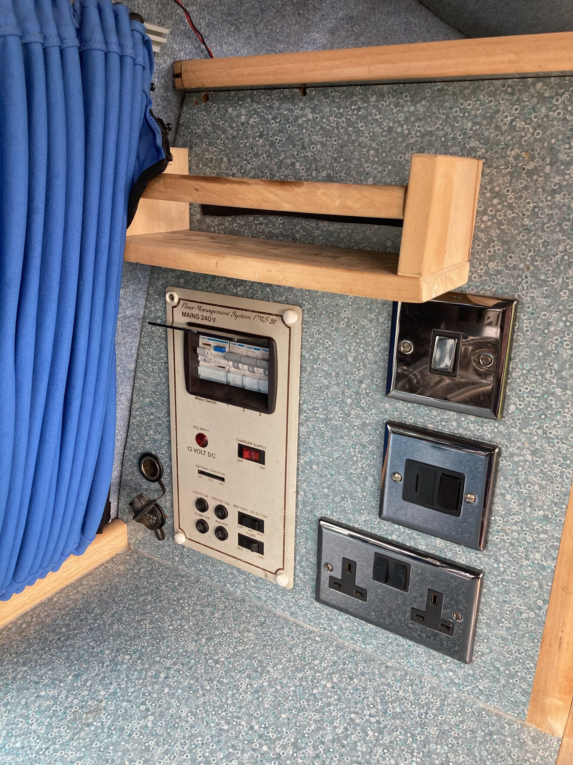 A close-up view of a wall panel in a caravan. The panel features multiple electrical outlets and switches, including UK-style power sockets, a light switch, a 12V DC outlet, and circuit breakers. Above the panel is a wooden rail and a blue curtain. The wall has a textured, light blue finish.
