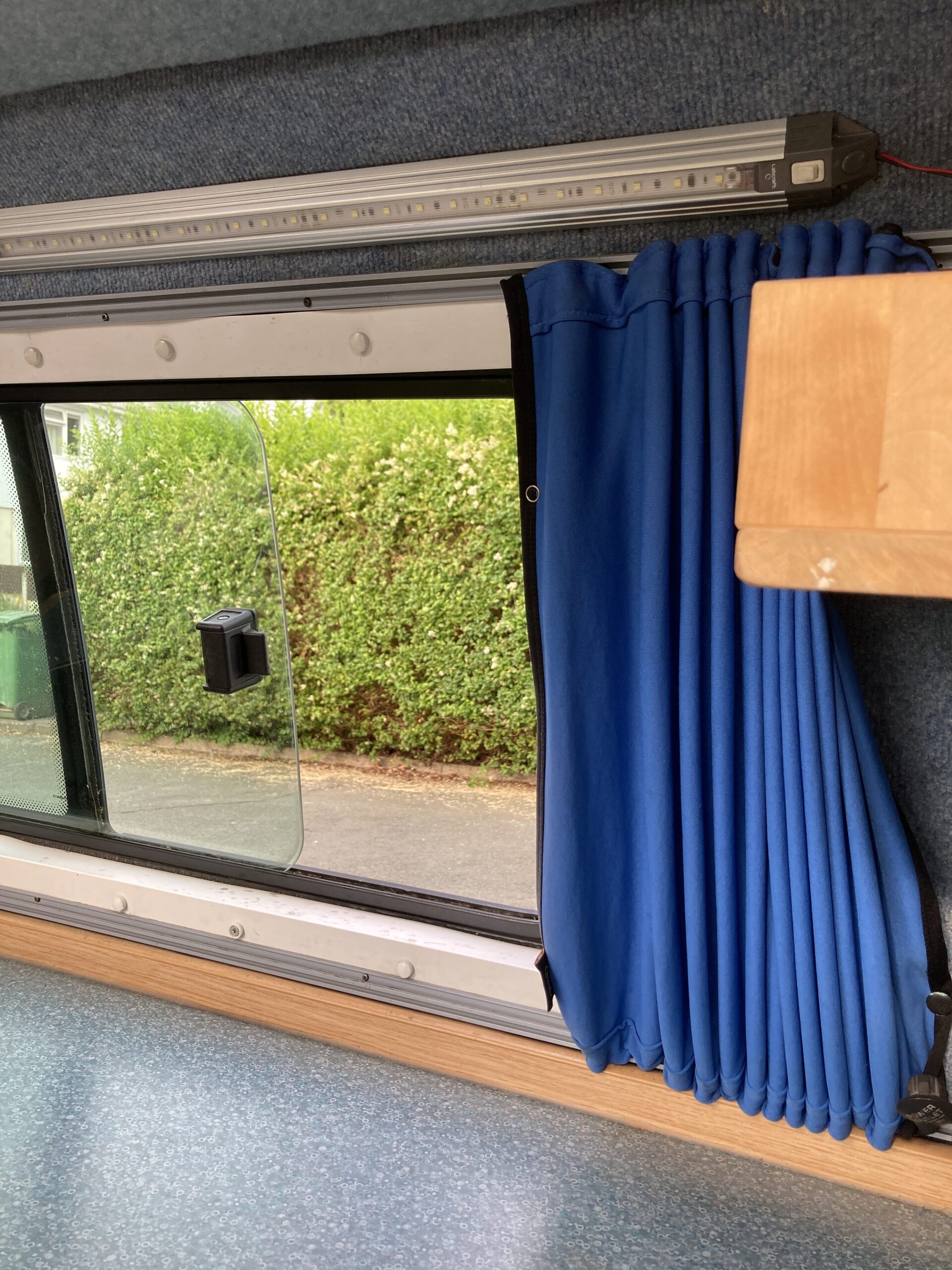 The interior view of a camper van shows a partially open window framed by a blue curtain gathered to the right. The window provides a view of vibrant green hedges outside. Above the window, a strip light is fixed, and a small wooden shelf is visible to the right side. The floor is covered in blue speckled linoleum.