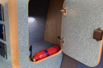 A small, open cabinet inside a vehicle, revealing a red fire extinguisher. The cabinet has a wood frame and door with teal speckled interior and sides. Adjacent is a cushioned seat with blue and gray fabric. The scene appears to be taken inside a camper van or RV.