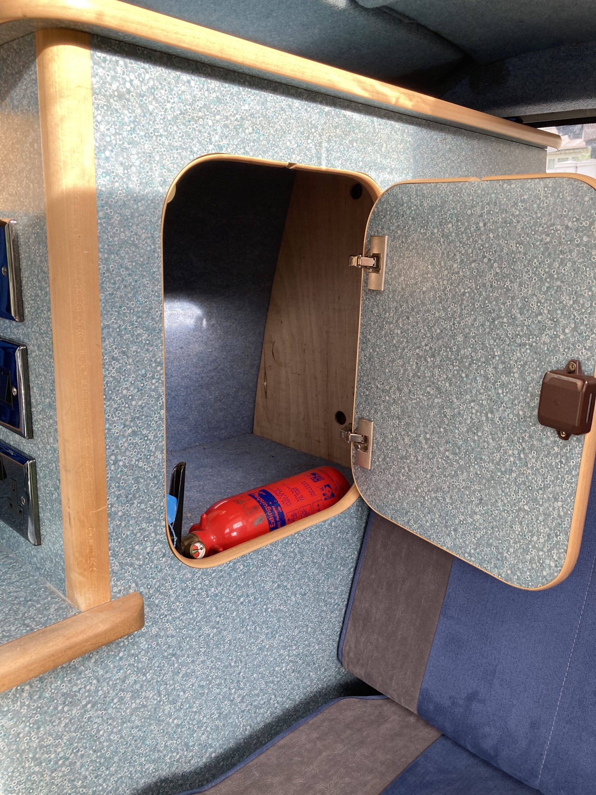 A small, open cabinet inside a vehicle, revealing a red fire extinguisher. The cabinet has a wood frame and door with teal speckled interior and sides. Adjacent is a cushioned seat with blue and gray fabric. The scene appears to be taken inside a camper van or RV.