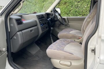 Interior view of an older vehicle with gray and beige fabric seats, a black rubber floor mat, and a black dashboard. The driver's door is open, showing manual controls and a simple, utilitarian design. The seating area is slightly worn, and the gear shift is visible between the seats.