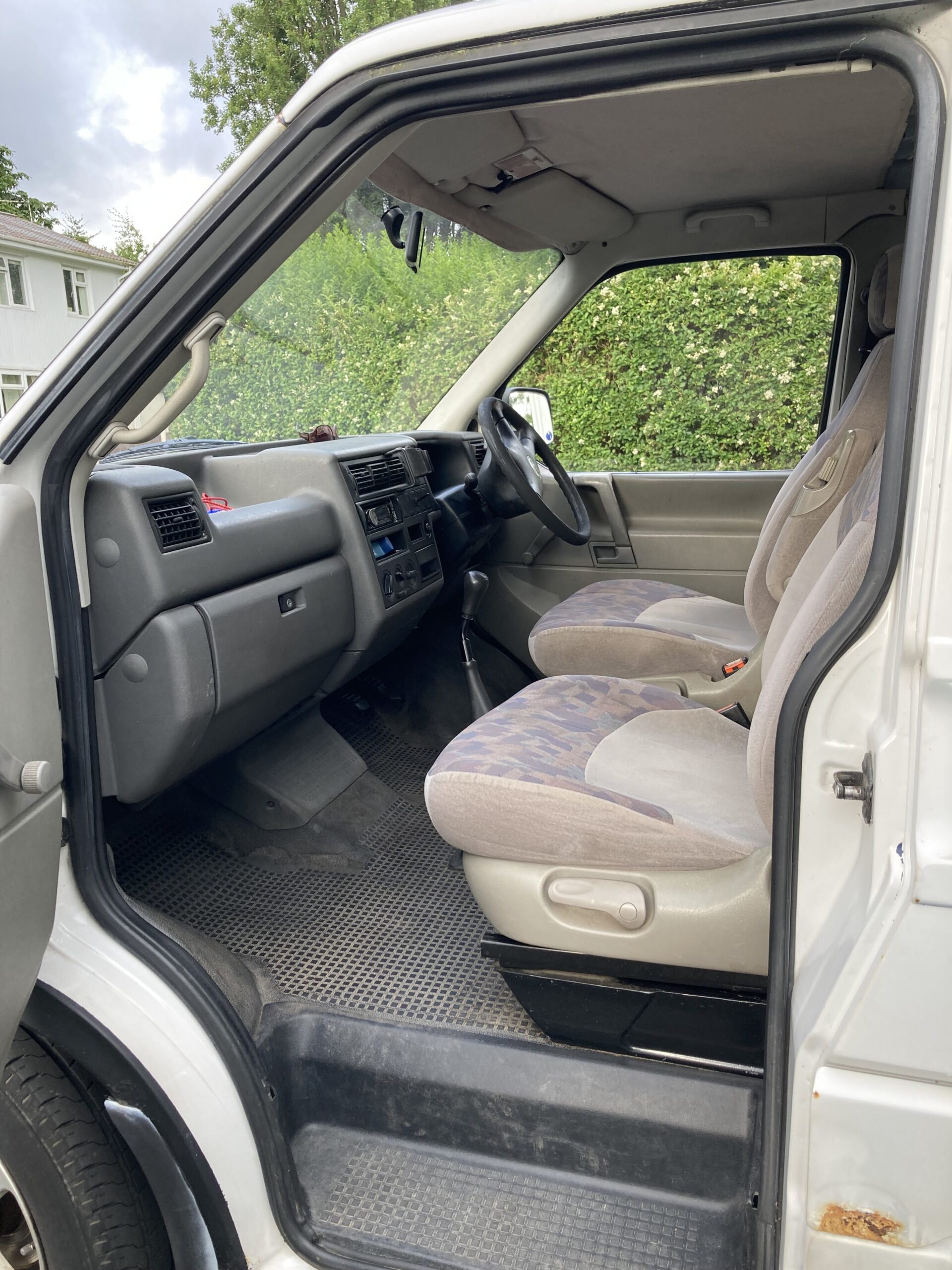 Interior view of an older vehicle with gray and beige fabric seats, a black rubber floor mat, and a black dashboard. The driver's door is open, showing manual controls and a simple, utilitarian design. The seating area is slightly worn, and the gear shift is visible between the seats.