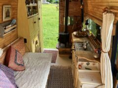 A cozy, wood-paneled camper interior features a small bed with patterned pillows on the left, a compact kitchen area with a farmhouse sink on the right, and a wood-burning stove at the far end. Natural light enters through a window above the sink, with a curtain partially drawn to one side.