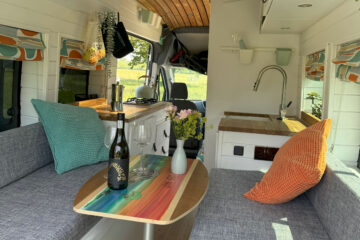 Interior of a stylish camper van with gray seating, colorful rainbow-striped table, and wooden ceiling. A bottle of wine, glasses, and a vase of flowers are on the table. Green and orange cushions add color, while a skylight brings in natural light. The rear door opens to a green grassy field.