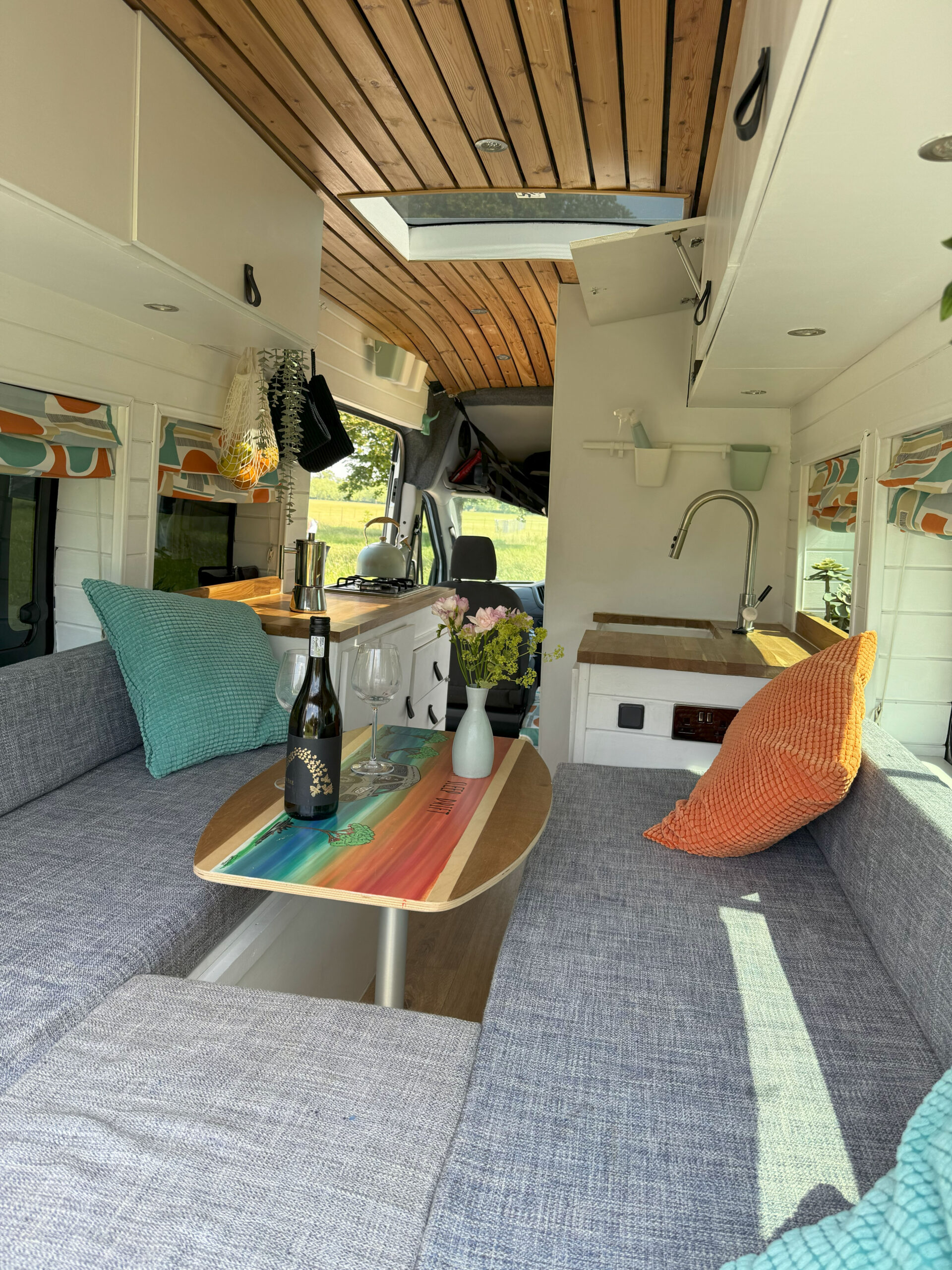 Interior of a stylish camper van with gray seating, colorful rainbow-striped table, and wooden ceiling. A bottle of wine, glasses, and a vase of flowers are on the table. Green and orange cushions add color, while a skylight brings in natural light. The rear door opens to a green grassy field.