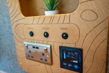 A wooden panel with intricate carvings features three labeled black buttons: "Water," "Front," and "Rear." Below them, there are two electrical outlets and a digital display showing "12.4V" and "23.5C." A small potted plant is placed in a shelf above the buttons.