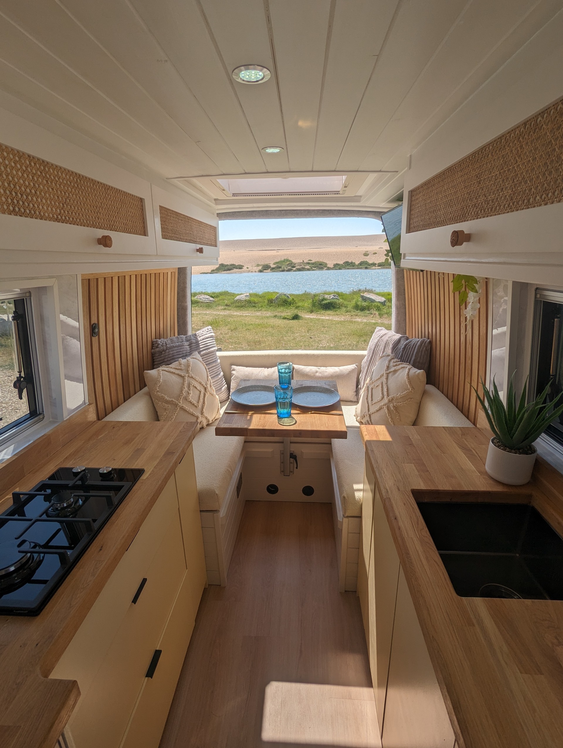 Interior of a cozy camper van with a modern design. The van features a small kitchen with a sink and stove, light wood countertops, and beige cabinets. A dining area at the back with a wooden table sits between cushioned benches with white pillows. Large windows offer a view of a lake and hills.