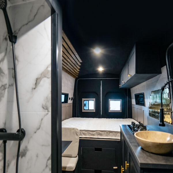 Interior of a modern, compact RV showcasing a luxurious design. Left foreground features a sleek showerhead and marble wall. Midsection has a cozy bed with two small windows above it. To the right, there's a kitchen area with a sink, stove, and minimalist cabinetry, all with stylish finishes.