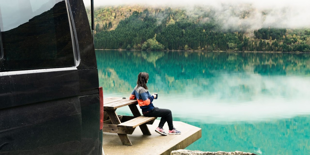 A person in a colourful jacket sits on a wooden picnic bench by the serene turquoise waters, holding a mug. A black van, known as the 83, is partially visible on the left. Across the lake, a dense forest and mist-covered mountains create a tranquil backdrop, highlighting one of the best wild swimming spots in the UK for 2024.