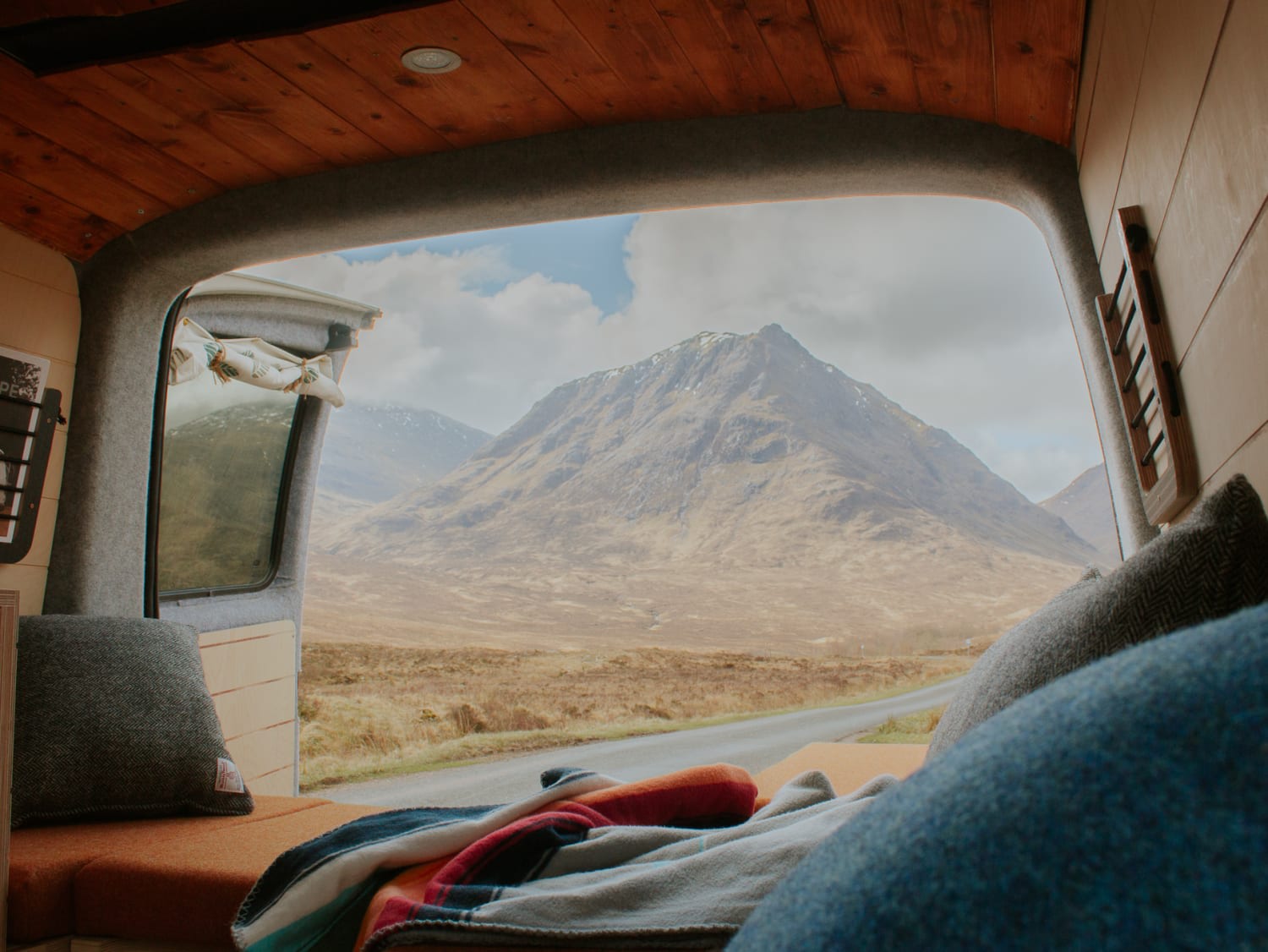 Inside a cosy campervan with a wooden interior, you can enjoy a scenic landscape featuring a large, rugged mountain under a cloudy blue sky. Blankets and cushions near the open door offer comfort and relaxation—the ideal setting for your jjppxtwcicfo6k8n87ng hire adventure away from Glasgow's bustling streets.